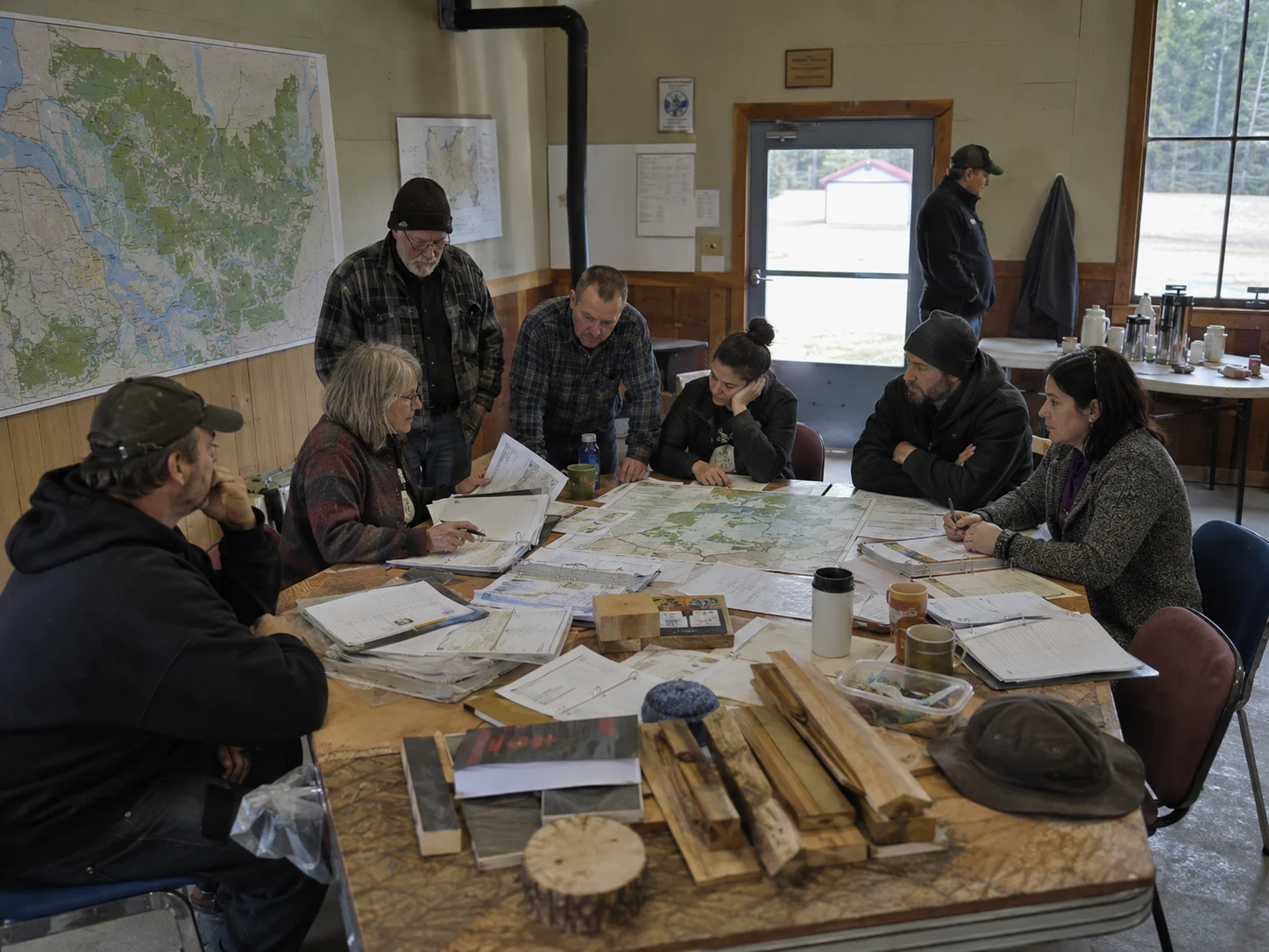 A modest public room where officials, builders, foresters, and community representatives work through maps, ledgers, timber samples, and documents.